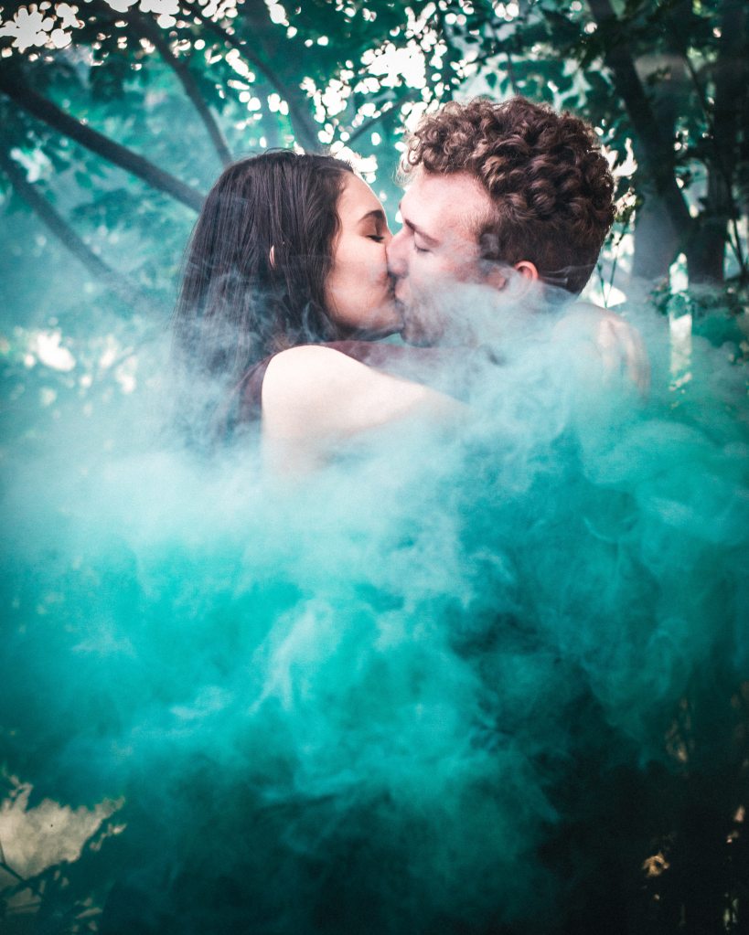 A photo of a man and a woman kissing in a forest as green and black smoke burst out before them, hiding most of their bodies from view