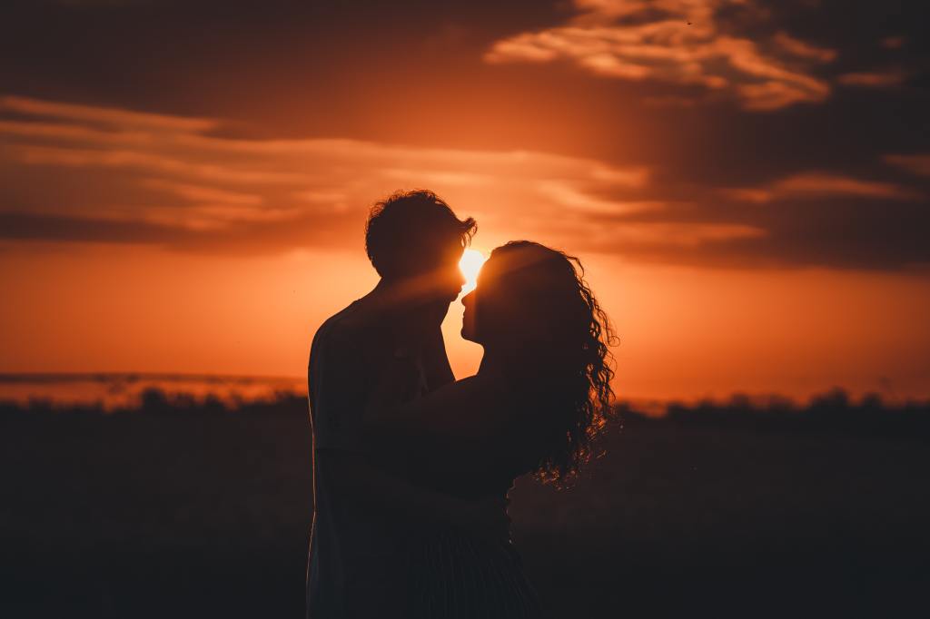 A photo of a man and a woman standing before a sunset. They are in silhouette and the setting sun is peeking out between them 